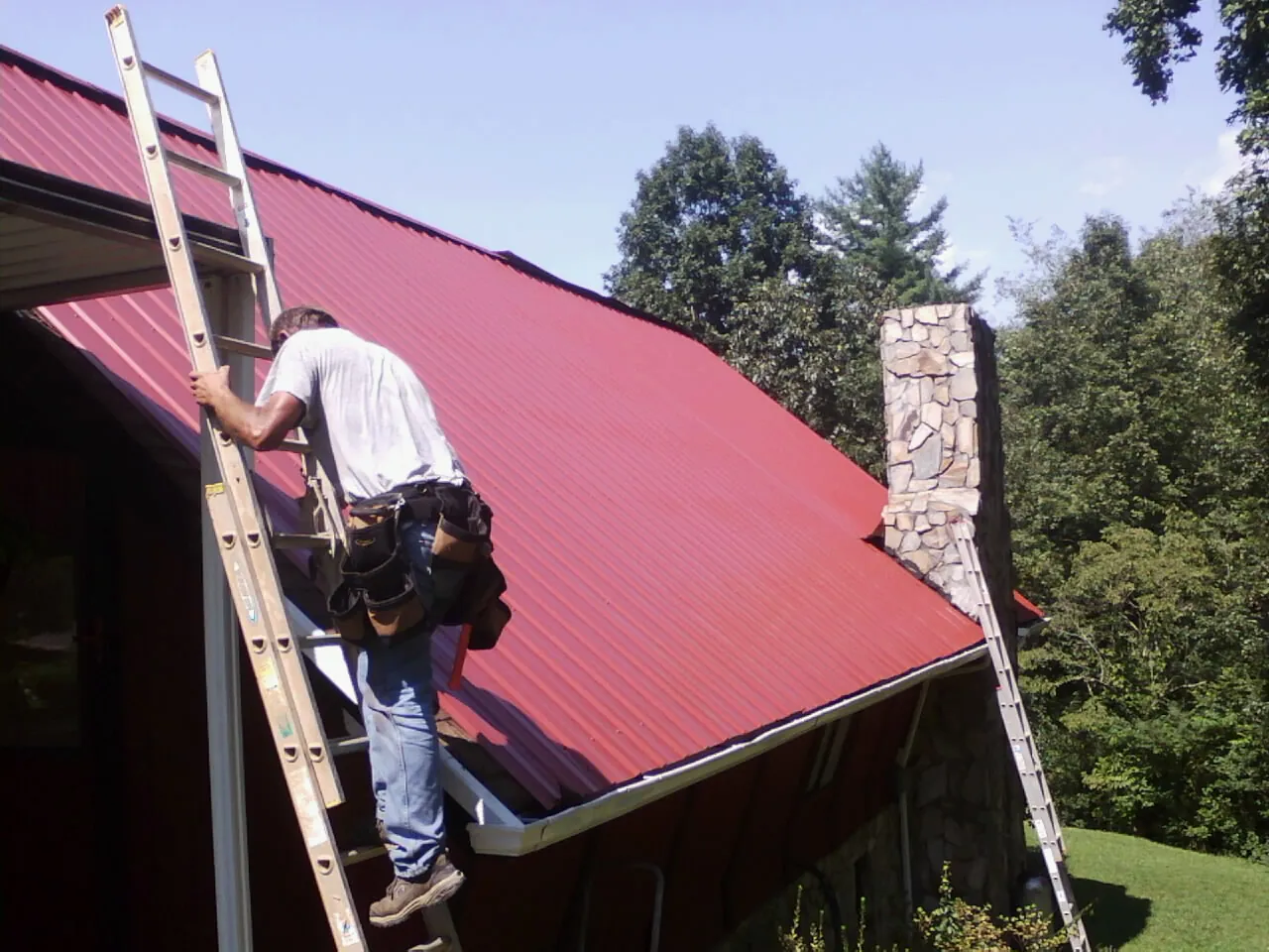 Roofer on a ladder beside a red metal roof and stone chimney — Soffit & Fascia Repair by Roofing Brothers in Spring House